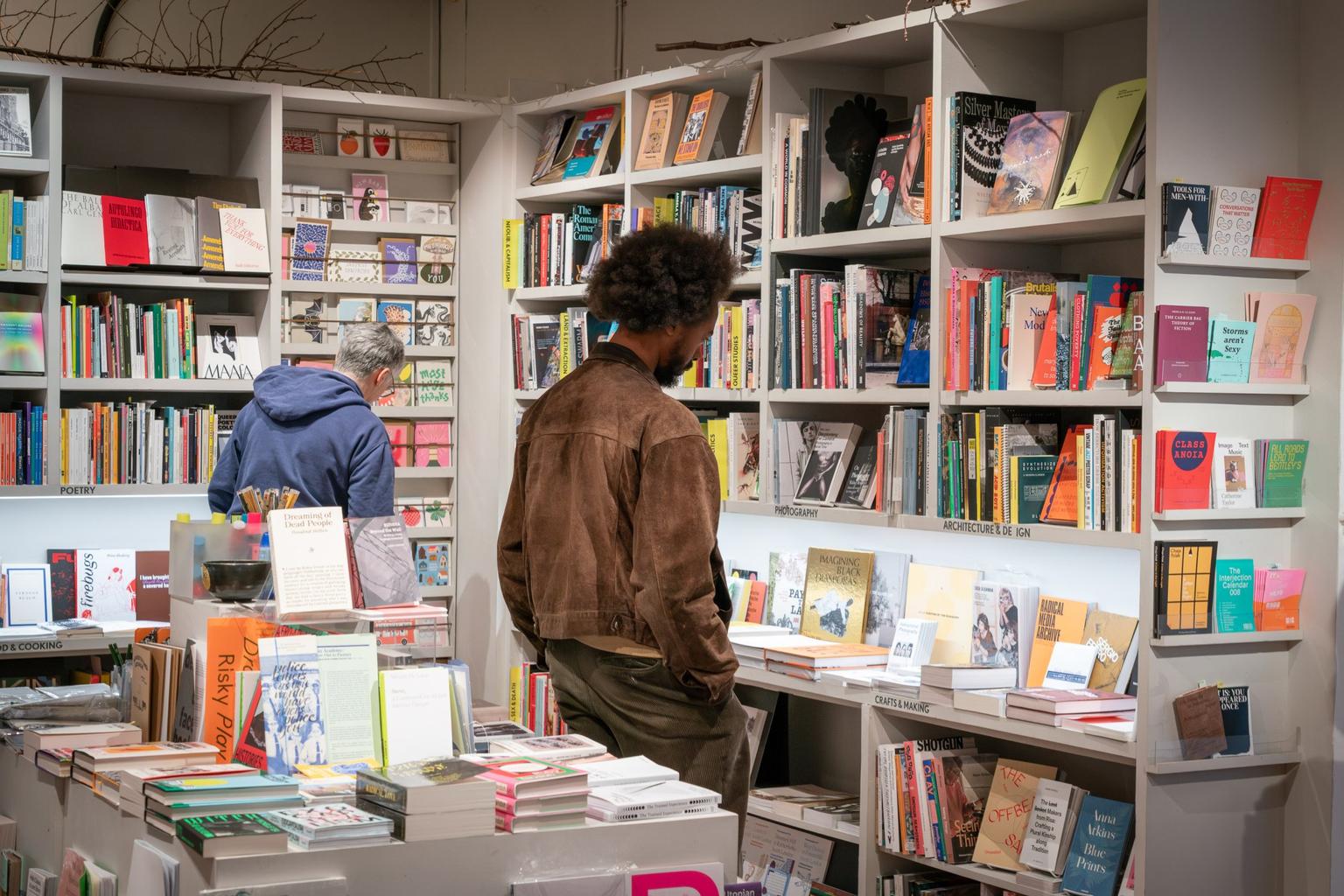 A person in a brown jacket browses books on white shelves in a brightly lit bookstore.