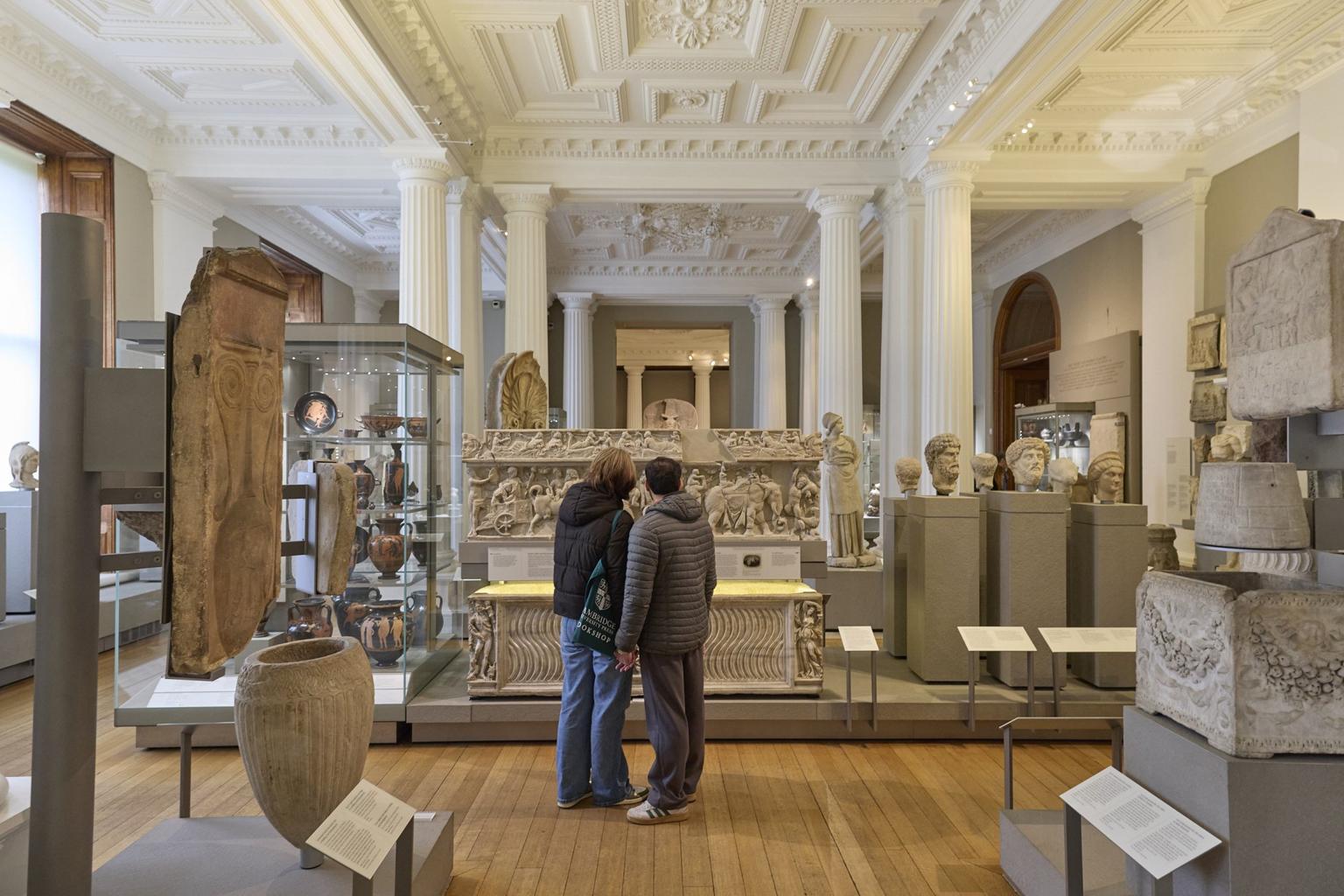Two visitors stand in a gallery of Greek and Roman art and archaeology.
