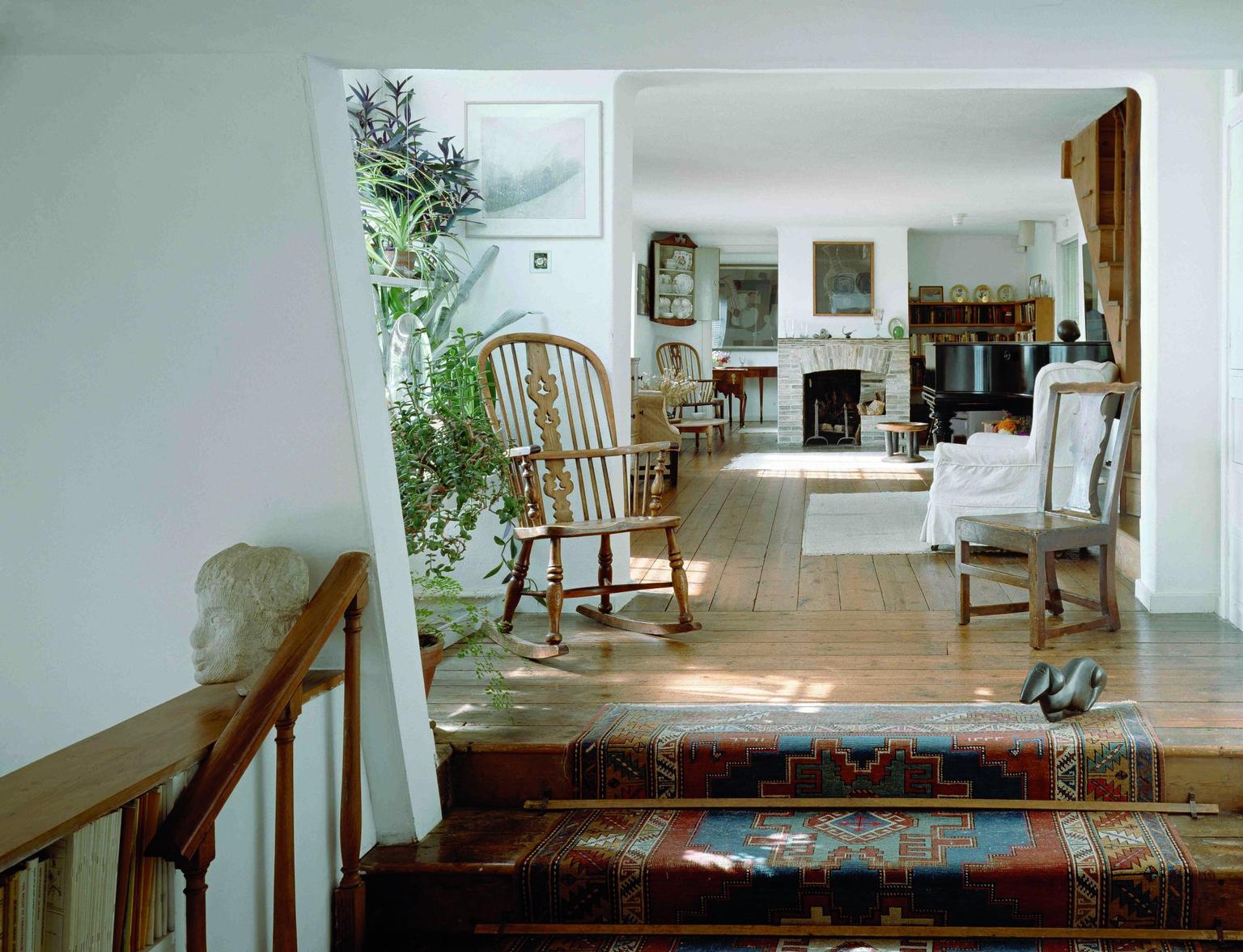 Sunlit living room with a wooden rocking chair, patterned red and blue stair runner in the foreground, a piano and stone fireplace visible beyond.