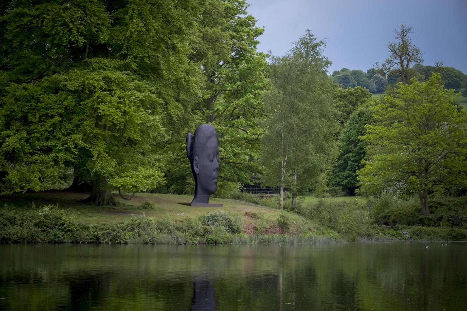 A huge black sculpture of a woman's head on a lush riverbank with mature trees