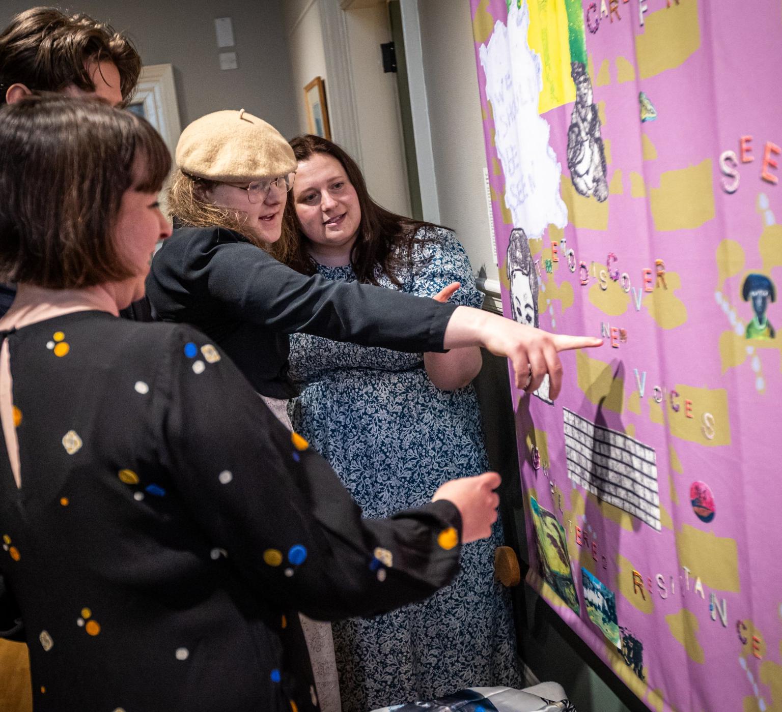 A small group of people stand close together pointing and discussing a bright pink and yellow collage-style banner covered in letters and images.
