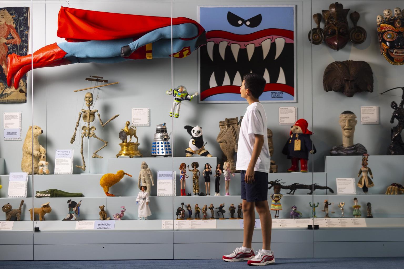 A boy looks into a glass cabinet, displaying figurines, teddies and toys, including Buzz Lightyear, Paddington Bear, Superman, Spice Girls Barbie dolls, Kung Fu Panda, and more.