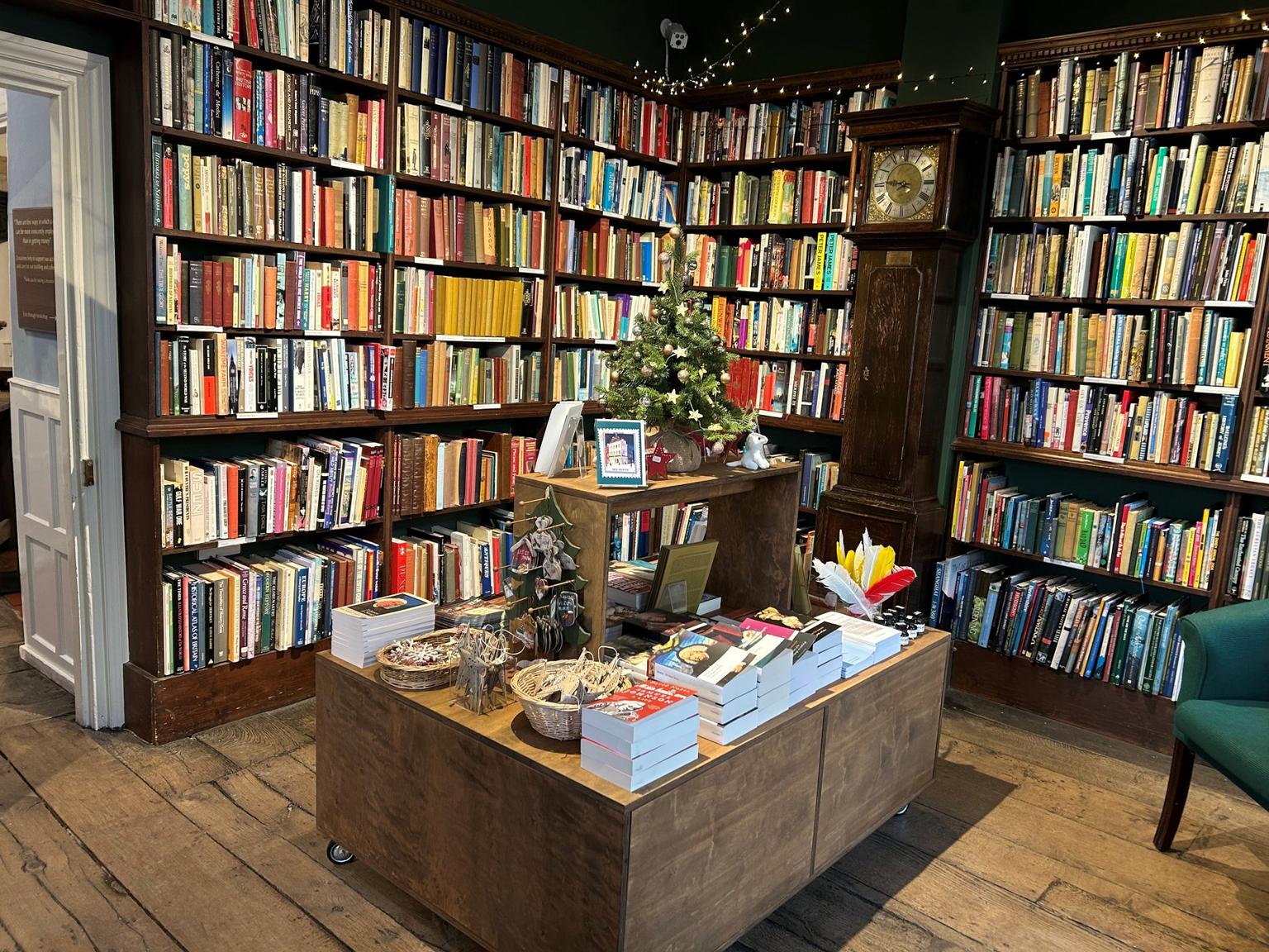 A cozy bookshop corner features tall wooden shelves filled with colorful books and a central display topped by a small Christmas tree.