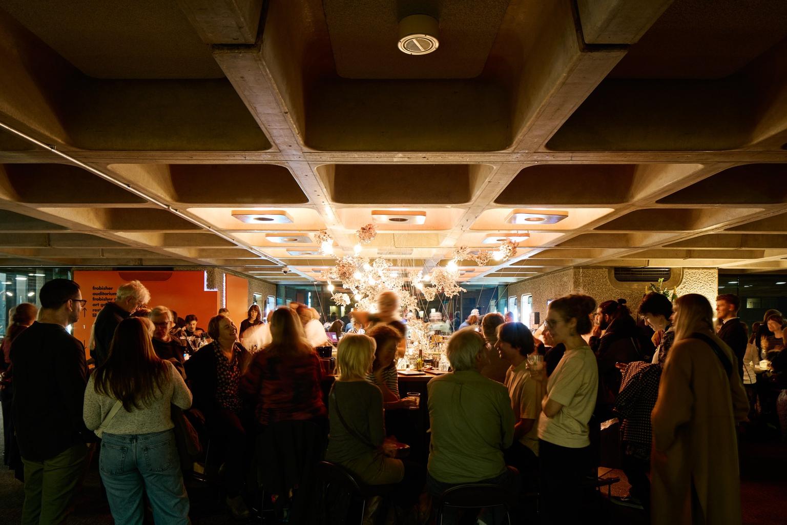 A crowded bar with a concrete ceiling and warm overhead lighting.