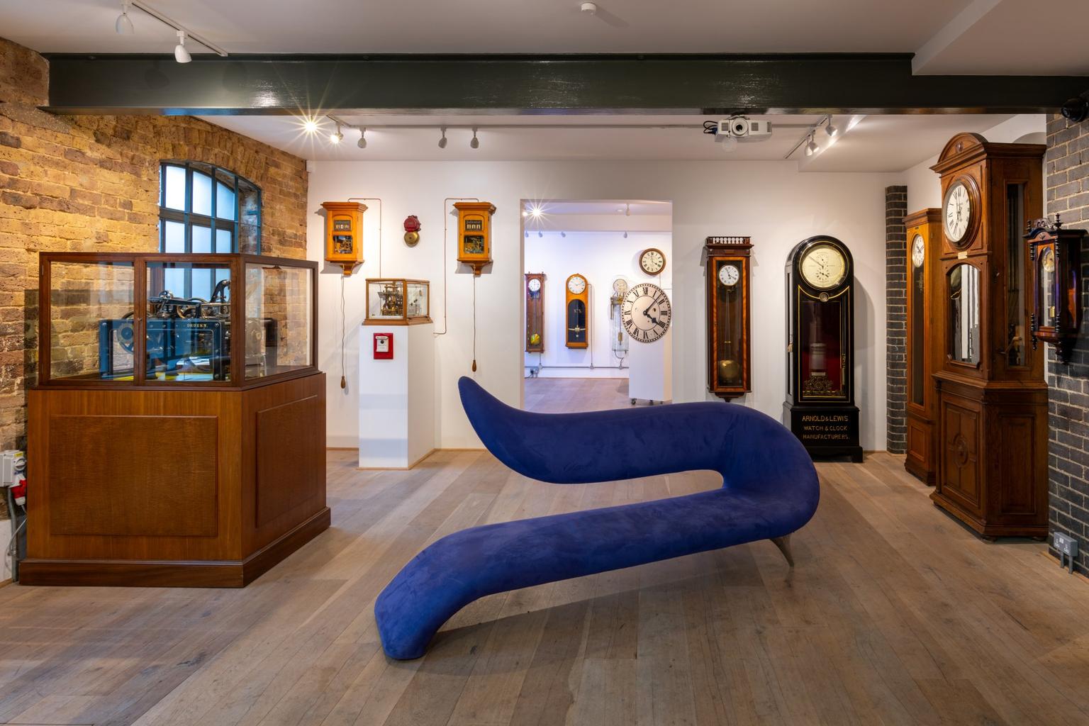 A curving cobalt-blue sculptural bench sits in the center of a museum room surrounded by antique wall and grandfather clocks.