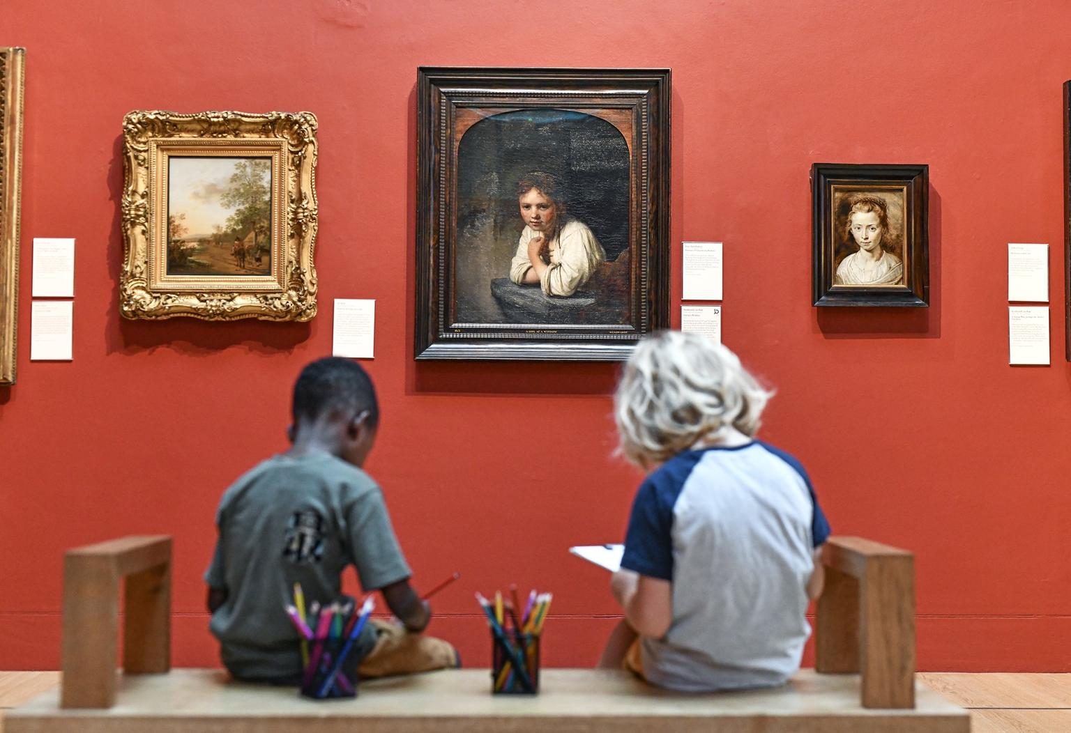 Two children on drawing while sitting on a bench in front of framed paintings on a red wall