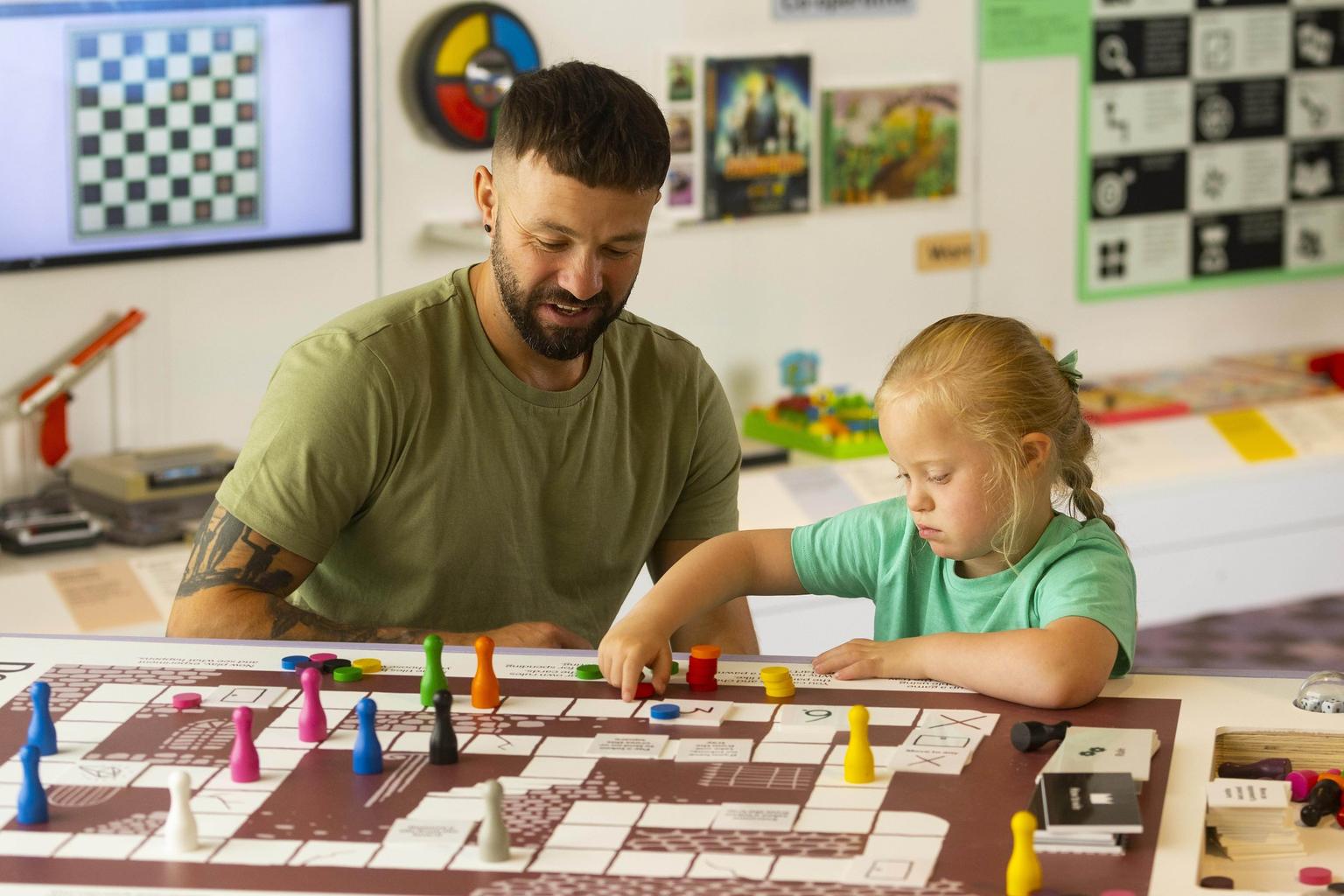 A young girl in a mint green T-shirt plays a board game with her father, who wears a dark green T-shirt.