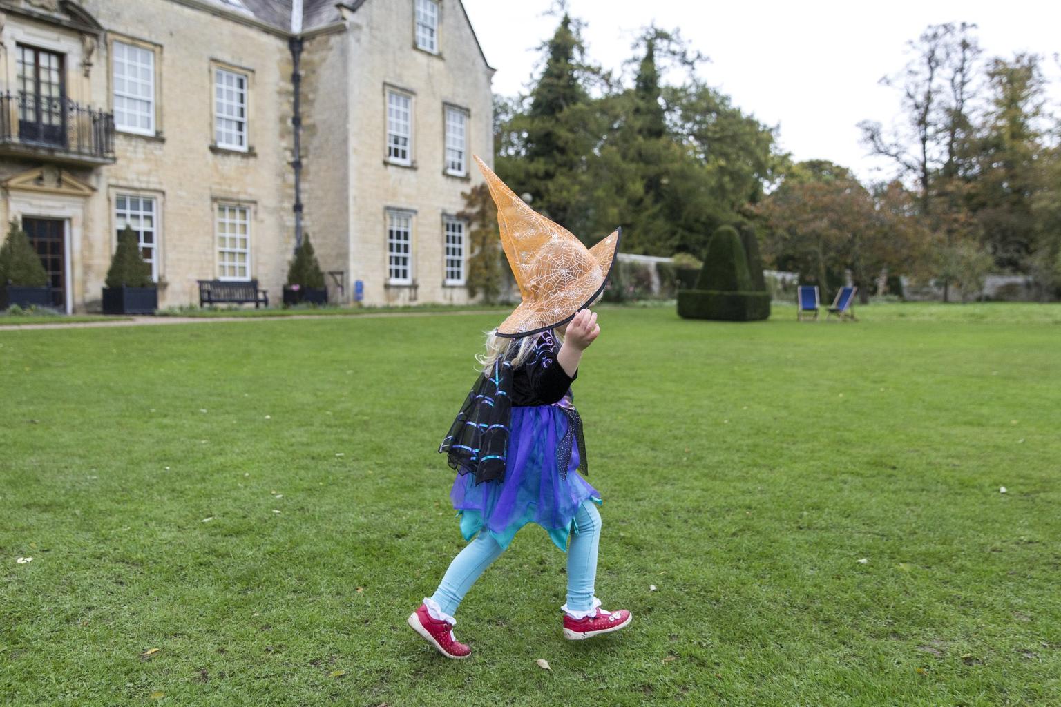A child in a bright orange witch hat and blue, purple and black costume runs across a lawn in front of a historic building.