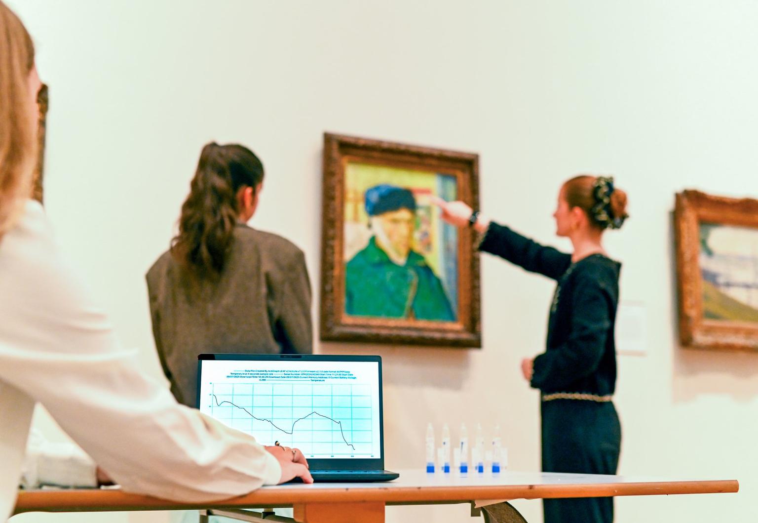Two visitors study a framed portrait in a bright gallery while a laptop on a nearby table displays a blue line chart.